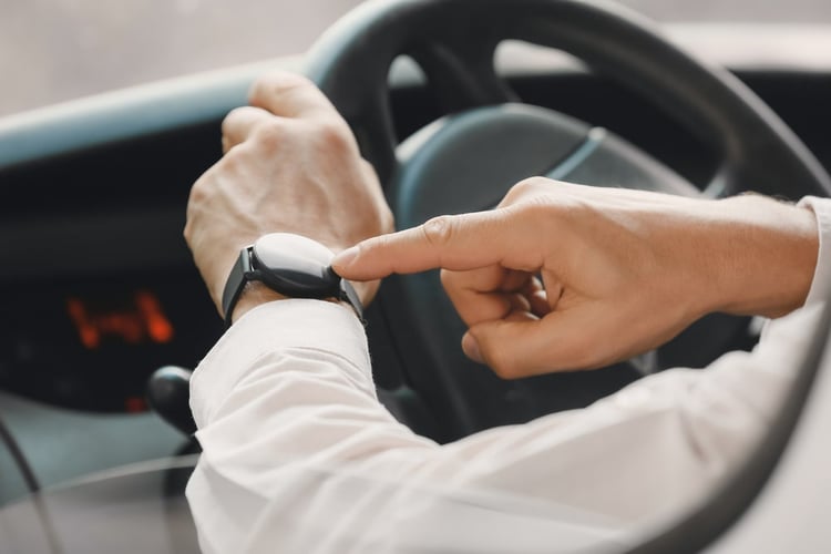 Un plan agrandi de 2 mains d'un homme au volant d'une voiture garée qui pointe vers sa montre.