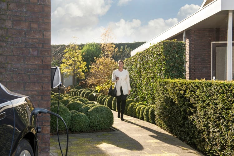 A woman walking through her well-kept garden, seemingly on her way to her electric car that's connected to an EVBox Elvi home charging station, mounted on the wall of her house.