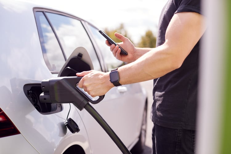 A man inserting a cable from the fast charger to his electric car while holding his smartphone in the other hand.