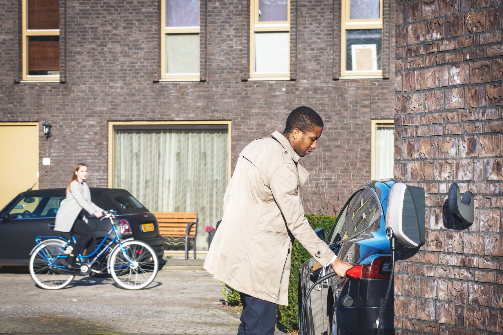 A casually dressed man plugging in his electric car outside a residential building on a sunny day. A woman who is biking in the background is looking at the man.