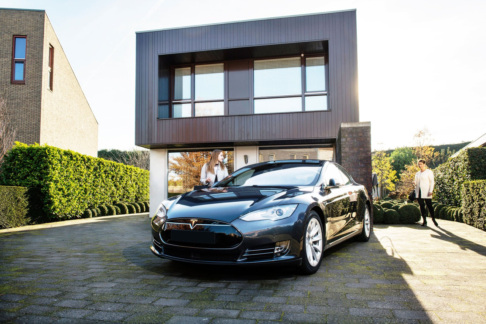 A black Tesla is parked in front of a modern house on a sunny day. A woman is approaching the vehicle and another person is standing in the background, next to the house.