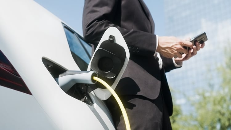 Man in black suit charging his electric car, leaning against it, with his smartphone in his hands.