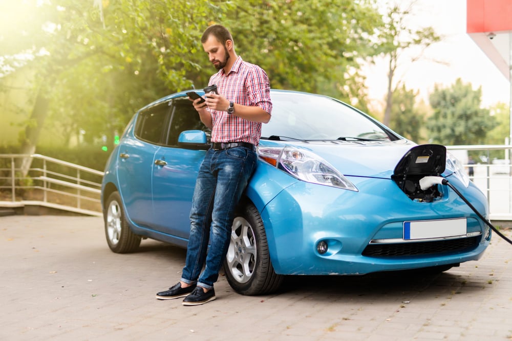 A man is checking his smart phone while leaning on his electric car whilst it is charging.