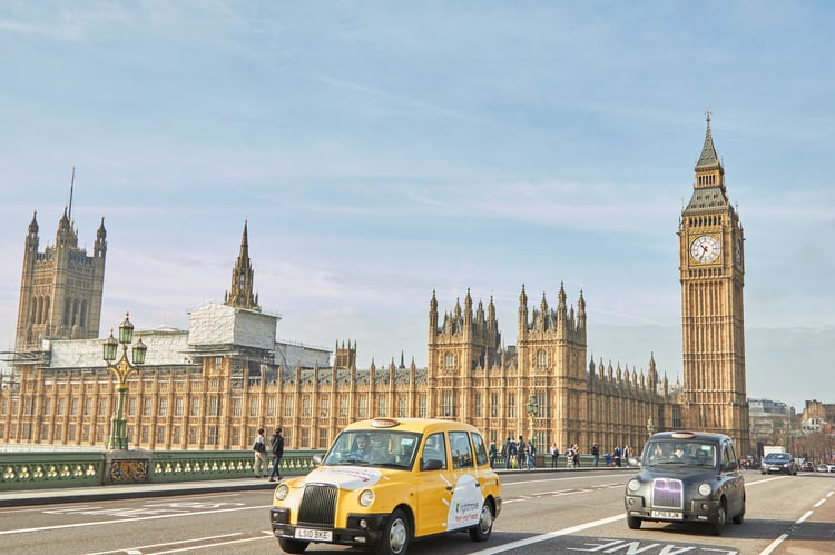 A yellow and black taxi driving past the houses of parliament on London's Westminster Bridge on a sunny day.