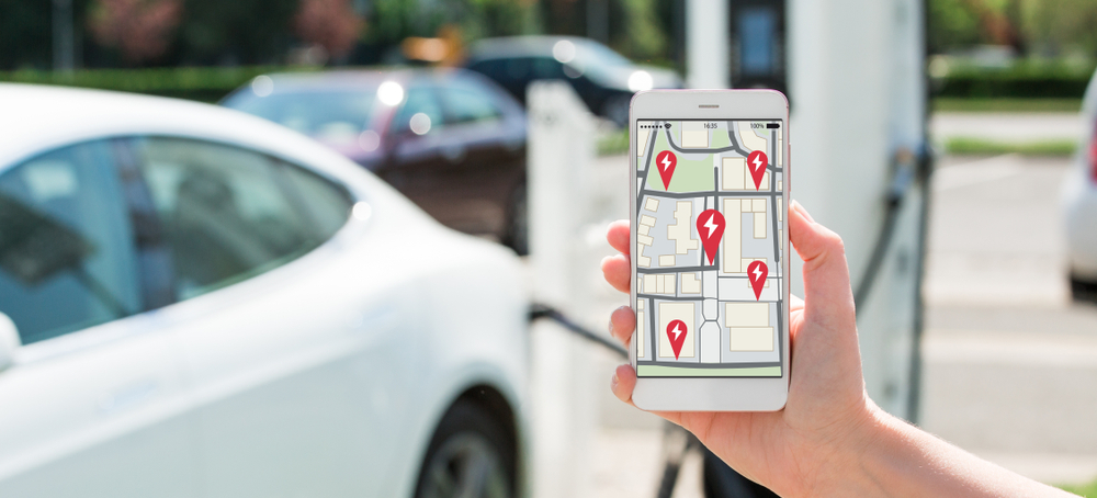 A hand holding a smart phone that displays an ev charging map. There is an electric car charging in the background.