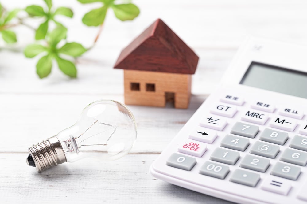 A closeup of a calculator, a light bulb and a small house model on a white wooden table.