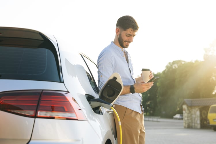 A smiling man is looking at his smartphone while checking EV charging data and insights of his electric car.