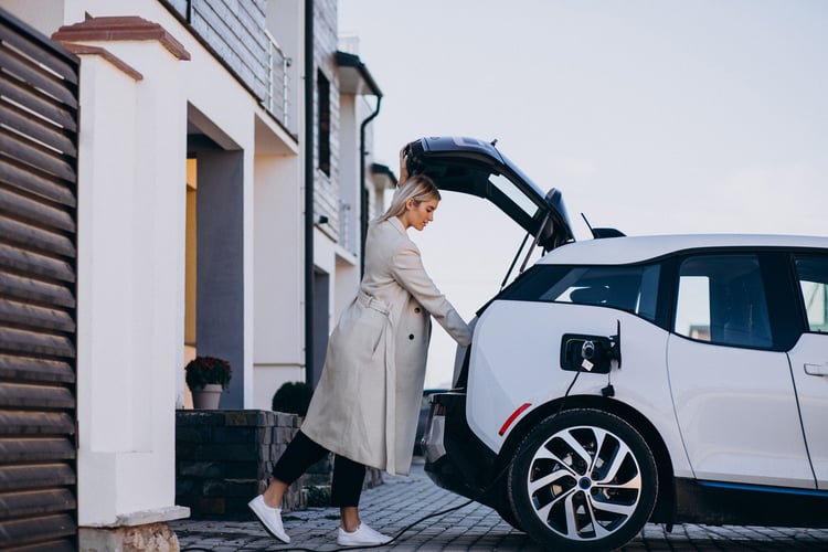 A blonde woman wearing a fashionable raincoat unloading her electric car whilst it’s charging in front of her house